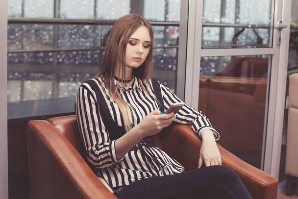 Young beautiful woman sits in a chair and looks in the phone, social network, chatting online. Wait your turn. Shopping center. The girl is dressed in a gray white striped blouse and corset. - Foto, Imagem