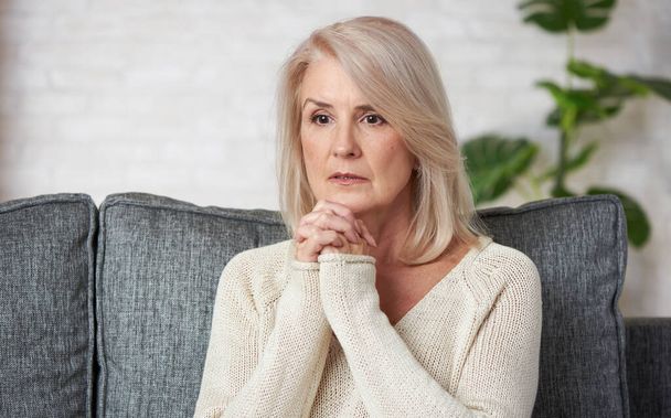 Portrait of sad mature woman sitting on couch at home and looking away with worry and anxiety - Photo, image