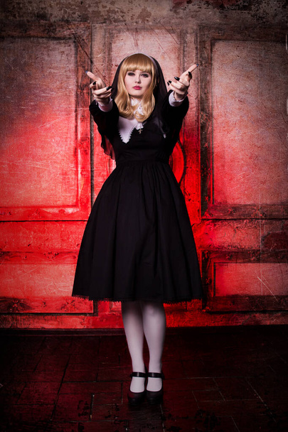 Religious nun in the concept of religion on a red background. Blonde with red eyes and dark makeup, vampire, demon. Costume for Halloween - Фото, изображение