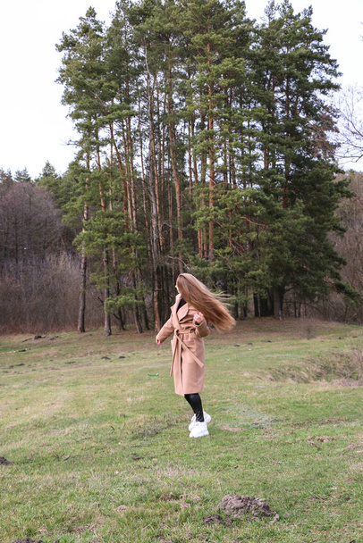 beautiful girl, blonde with long hair, walks in the spring near the forest in a brown coat., serenity and freedom - Foto, Bild