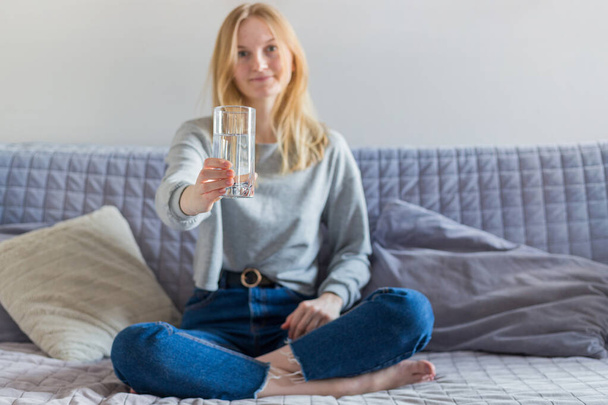 Pretty blonde woman drinking water and sitting on the sofa at home in the living room. Beautiful young girl with glass of water - Fotoğraf, Görsel