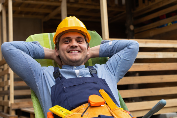 Picture of happy young carpenter with uniform and yellow helmet resting after hard work outdoor - Foto, Imagen