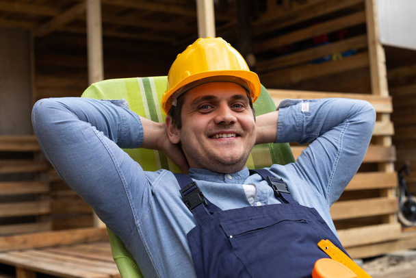 Picture of happy young carpenter with uniform and yellow helmet resting after hard work outdoor - Foto, Imagem