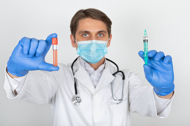 Young male doctor with surgical mask and gloves holding a blood sample and injection on white background - Foto, Imagen