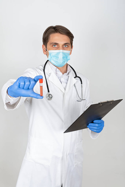 Young male doctor with surgical mask and gloves holding a blood sample on white background - Photo, image
