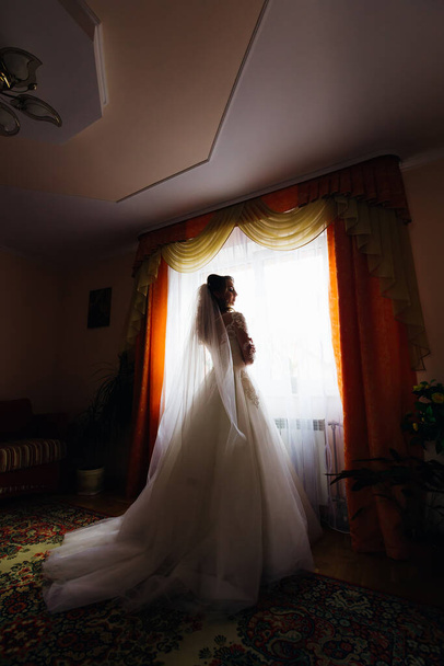 Die Braut im weißen Kleid mit Brautschleier steht am Fenster mit Vorhängen - Foto, Bild