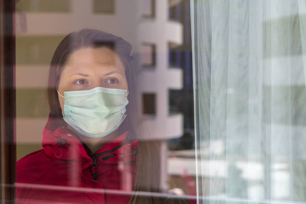 Girl or young woman in a medical mask being quarantined at home during the coronavirus pandemic. Self-isolation, view through glass, walking on the balcony of an apartment in city - Fotografie, Obrázek