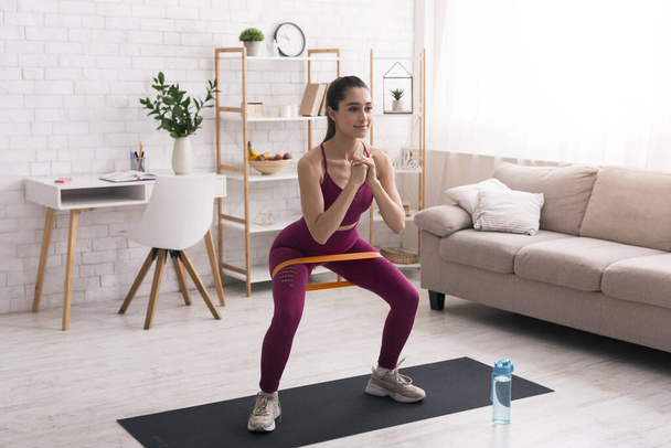 Thuis sporten. Spaans meisje doet squats met elastische band in de woonkamer - Foto, afbeelding