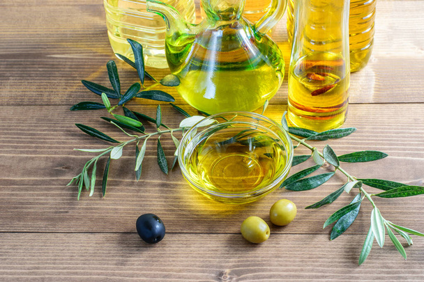 Ассортимент бутылок с оливковым маслом, оливковое масло в прозрачной стеклянной чаше, оливки и ветви оливкового дерева на деревянном фоне. Тяжелое питание
.  - Фото, изображение