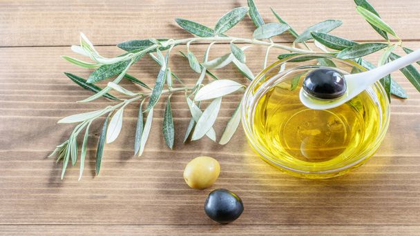 Olive dans une cuillère, huile d'olive goutte à goutte dans un bol transparent en verre. Quelques olives et des branches d'olivier sur le fond en bois
.  - Photo, image