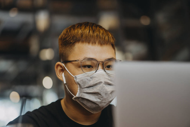 Asian young man indoors in cafe. Lifestyle concept photo with copy space. Picture with handsome guy who using protective face mask. Portrait with gray laptop - Photo, Image