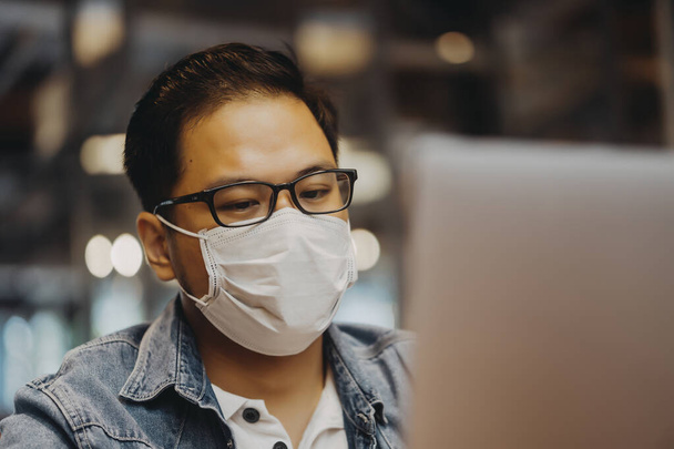 Asian young man indoors in cafe. Lifestyle concept photo with copy space. Picture with handsome guy who using protective face mask. Portrait with gray laptop - Foto, immagini