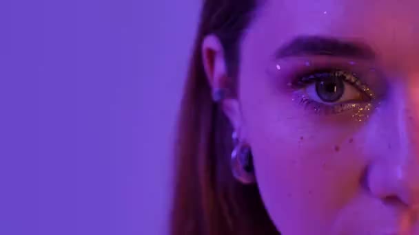 A cropped view of an eye of a woman with fashion glitter makeup is looking to the camera and winking isolated over violet neon background - Felvétel, videó