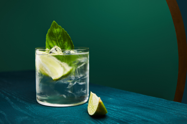 close up view of old fashioned glass with drink and lime on blue wooden surface isolated on green - Photo, Image