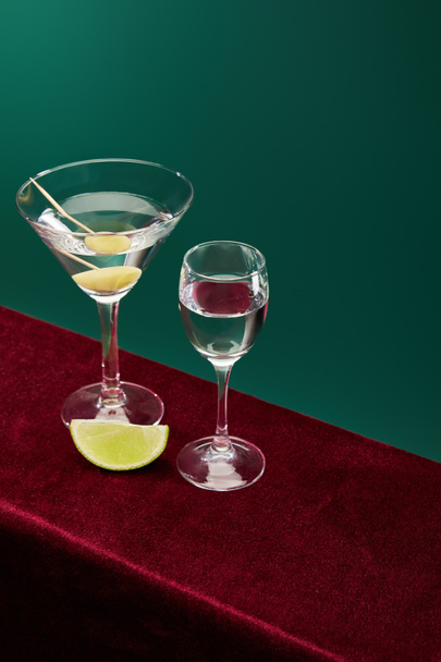 high angle view of shot glass and cocktail glass with vermouth, lime slice and whole olive on toothpick on red velour surface isolated on green - Photo, Image