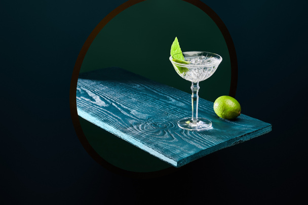 high angle view of cocktail glass with mint leaf and whole lime on blue wooden surface on geometric background - Photo, Image