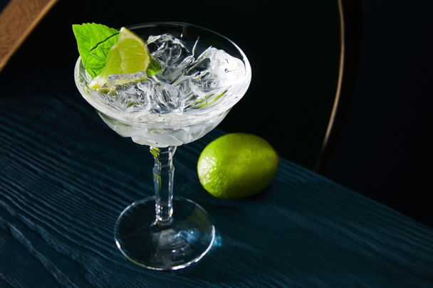 High angle view of cocktail glass with ice cubes, mint leaf and whole lime on blue wooden surface on geometric background - 写真・画像