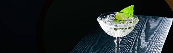high angle view of cocktail glass with ice cubes and mint leaf on blue wooden surface on black background - Фото, изображение