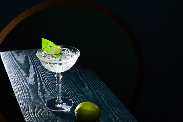 high angle view of cocktail glass with drink with ice cubes, mint leaf and whole lime on blue wooden surface on black background - Fotografie, Obrázek