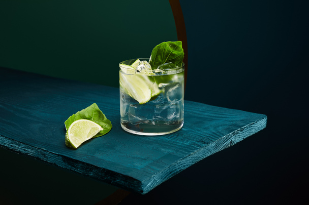old fashioned glass with fresh drink, mint leaf and lime slice on blue wooden surface on green and blue geometric background - Photo, Image