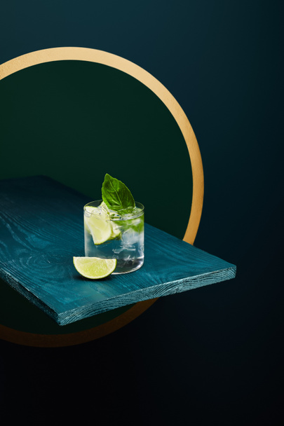 High angle view of old fashioned glass with fresh drink, mint leaf and lime slice on blue wooden surface on green and blue geometric background with golden circle - Photo, Image