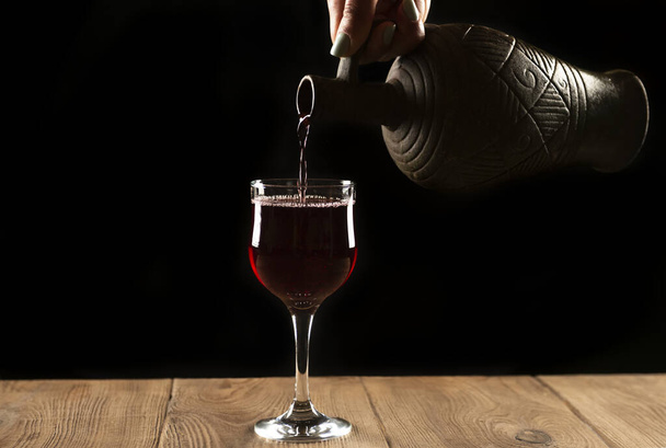girl hands pouring red wine into a glass from a clay bottle on a black background - Fotó, kép
