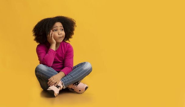 Pensive little girl sitting on floor over yellow background and thinking - Fotó, kép
