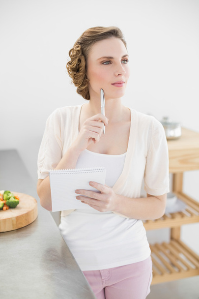 Thinking woman standing in kitchen writing a shopping list - Photo, image