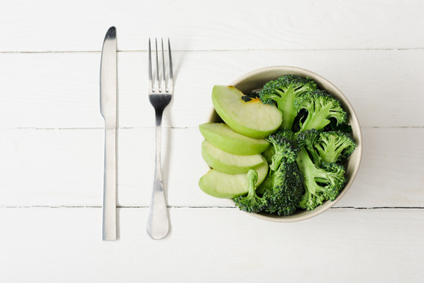 top view of fresh green apple and broccoli in bowl near cutlery on white wooden surface - Foto, Imagem