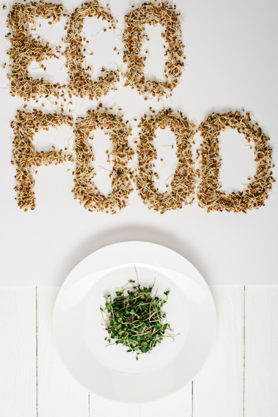 πάνω όψη του φρέσκου microgreen στο πιάτο κοντά σε οικολογικά γράμματα τροφίμων από λαχανάκια σε λευκή ξύλινη επιφάνεια - Φωτογραφία, εικόνα