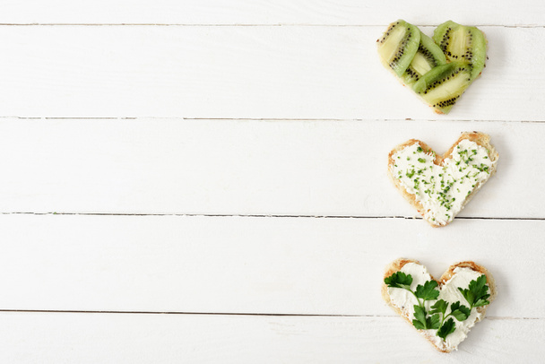 κορυφαία άποψη του canape σε σχήμα καρδιάς με κρεμώδες τυρί, μικροπράσινο, μαϊντανό και ακτινίδιο σε λευκή ξύλινη επιφάνεια - Φωτογραφία, εικόνα