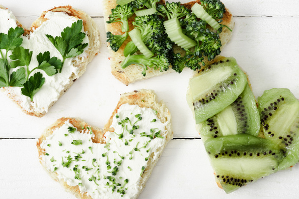 vrchní pohled na srdíčko ve tvaru canape s krémovým sýrem, brokolicí, microgreen, petrželkou a kiwi na bílém dřevěném povrchu - Fotografie, Obrázek