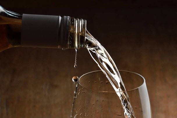 Weißwein wird ins Glas gegossen. Weinströme aus der Flasche und Wirbel und Blasen im Glas. Glas mit goldener Flüssigkeit auf Holztisch. - Foto, Bild