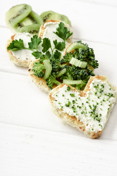 selective focus of heart shaped canape with creamy cheese, broccoli, microgreen, parsley and kiwi on white wooden surface - Foto, Imagen