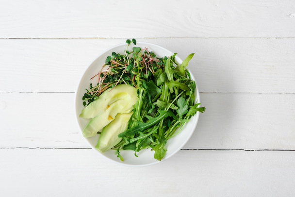 top view of arugula, avocado and microgreen in bowl on white wooden surface - Photo, Image