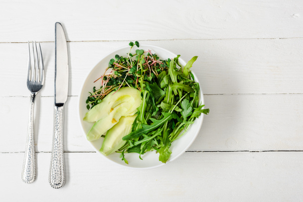 top view of arugula, avocado and microgreen in bowl on white wooden surface with cutlery - 写真・画像