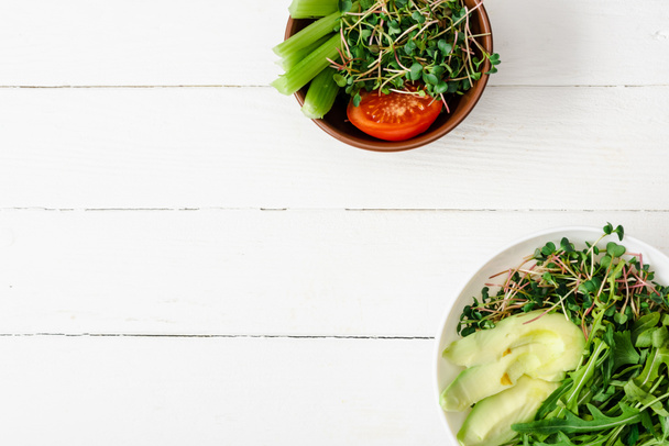 Draufsicht auf frisches Gemüse mit Avocado und Microgreen in Schalen auf weißer Holzoberfläche - Foto, Bild