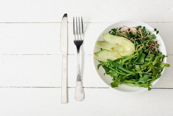 top view of arugula, avocado and microgreen in bowl on white wooden surface with fork and knife - Fotó, kép