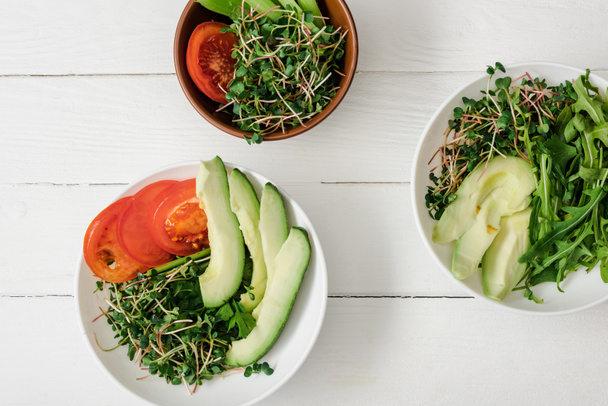 top view of fresh vegetables with avocado and microgreen in bowls on white wooden surface - 写真・画像