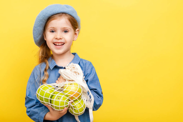 A little blonde girl with long hair in a blue dress and beret on a yellow isolated background. Child with apples. - Fotografie, Obrázek