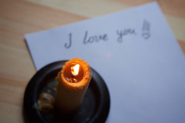 Η επιγραφή "Σ 'αγαπώ" με τη μορφή επιστολής με το χέρι σε λευκό χαρτί και ενός κεριού που καίει στο τραπέζι. Ερωτικό γράμμα. - Φωτογραφία, εικόνα