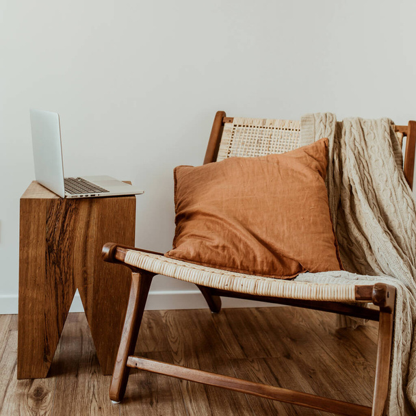 Concepto moderno de diseño de interiores. Elegante silla de madera de ratán, a cuadros de punto, almohada de jengibre. Acogedor cómodo espacio de trabajo de oficina en casa con computadora portátil
. - Foto, Imagen