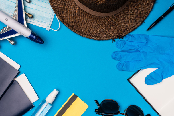 Top view of toy plane near medical mask, sun hat with passports and latex glove on blue background - Foto, afbeelding