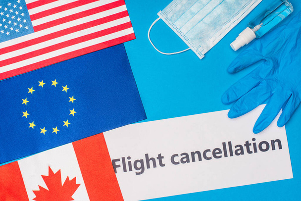 Top view of card with flight cancellation lettering near medical mask and flags of countries on blue surface - Photo, Image