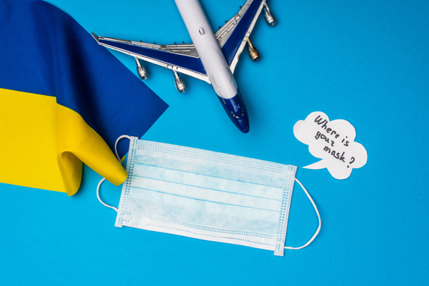 Top view of toy airplane near medical mask, flag of ukraine and speech bubble with where is your mask lettering on blue surface - Photo, Image