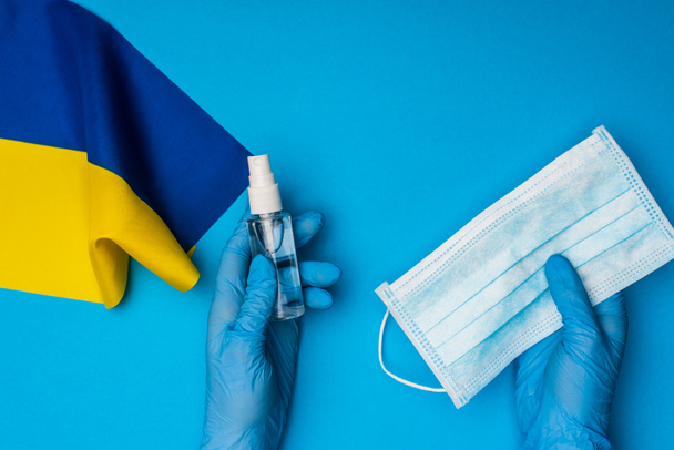 Top view of doctor holding bottle with hand sanitizer and medical mask near flag of ukraine on blue background - 写真・画像