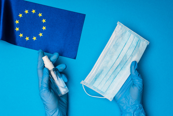 Top view of doctor holding medical mask and bottle of hand sanitizer near flag of european unity on blue background - Photo, Image