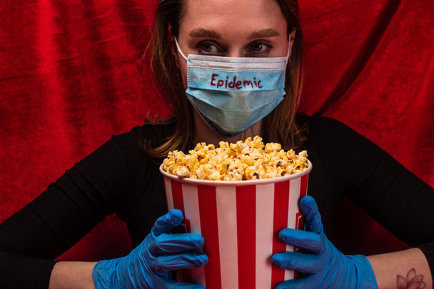 Woman in medical mask with epidemic lettering and latex gloves holding bucket with popcorn with red velour at background - Photo, image