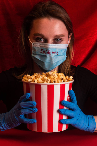 Młoda kobieta w masce medycznej z epidemiologicznym napisem trzymająca wiadro z popcornem na czerwonej powierzchni z welurowym tłem - Zdjęcie, obraz