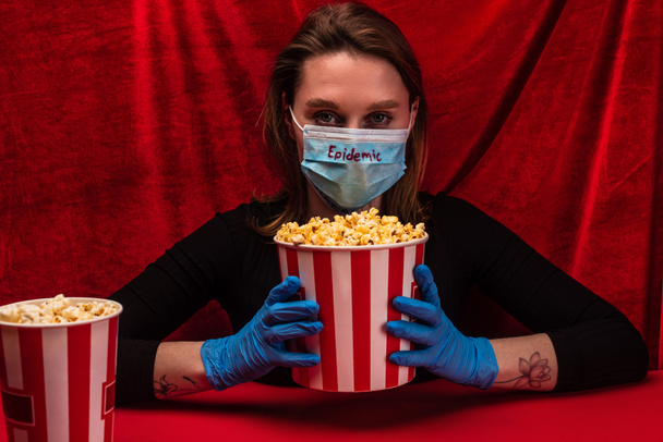 Woman in latex gloves and medical mask with epidemic lettering holding bucket with popcorn on red surface with velour at background - Zdjęcie, obraz